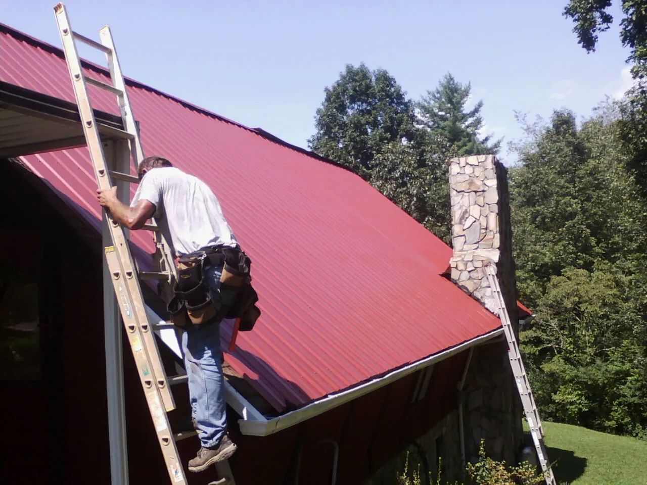 Roofer on a ladder beside a red metal roof and stone chimney — Soffit & Fascia Repair by Roofing Brothers in Ramona