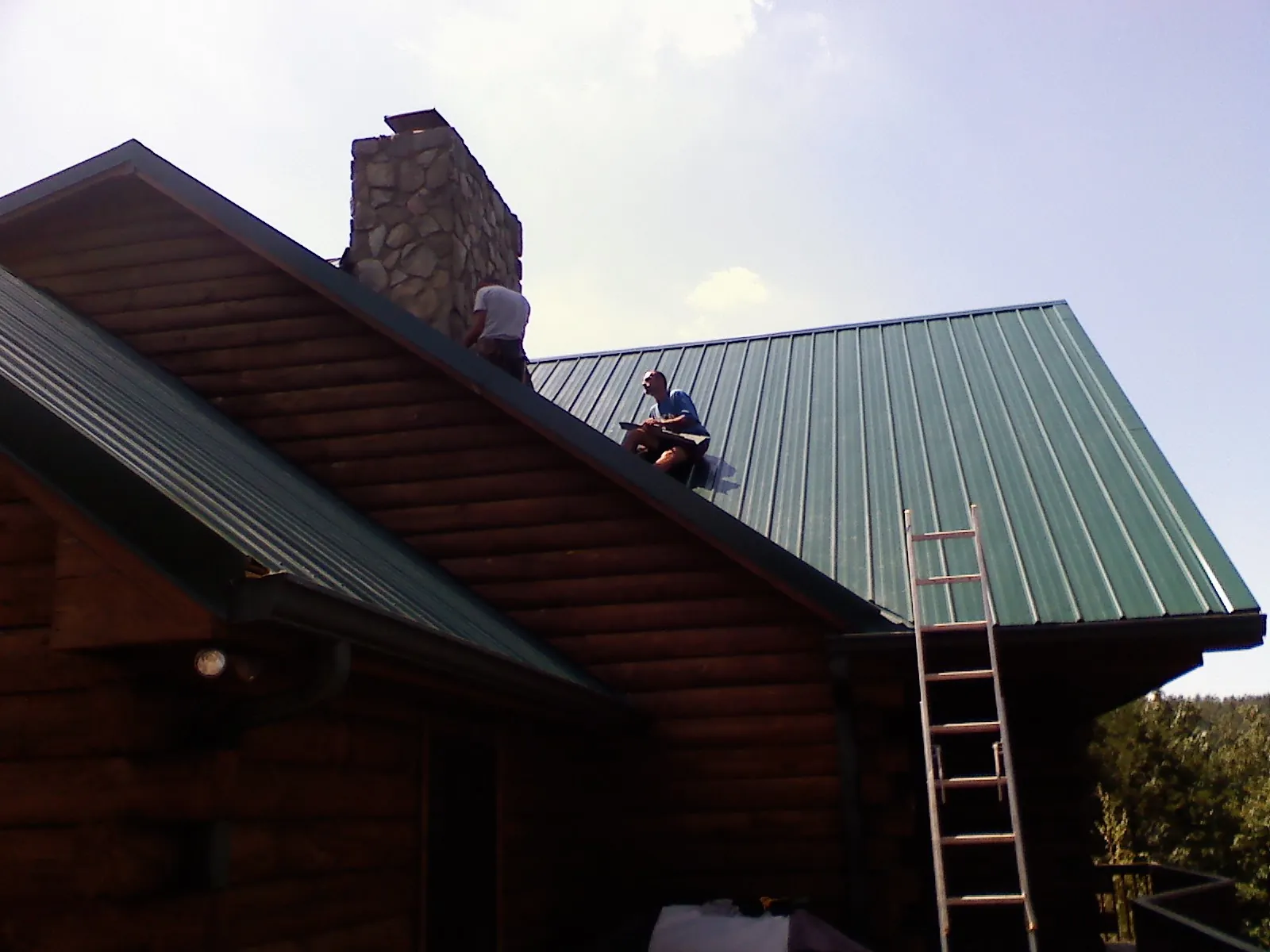 Roofers installing green metal panels beside a stone chimney on a log home in Ramona